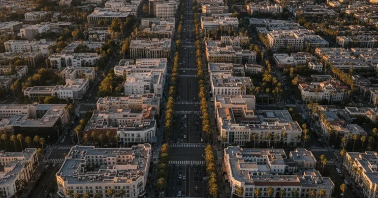Photorealistic wide-angle aerial view of Beverly Hills city skyline at golden hour, showing palm-tree-lined boulevards, luxur